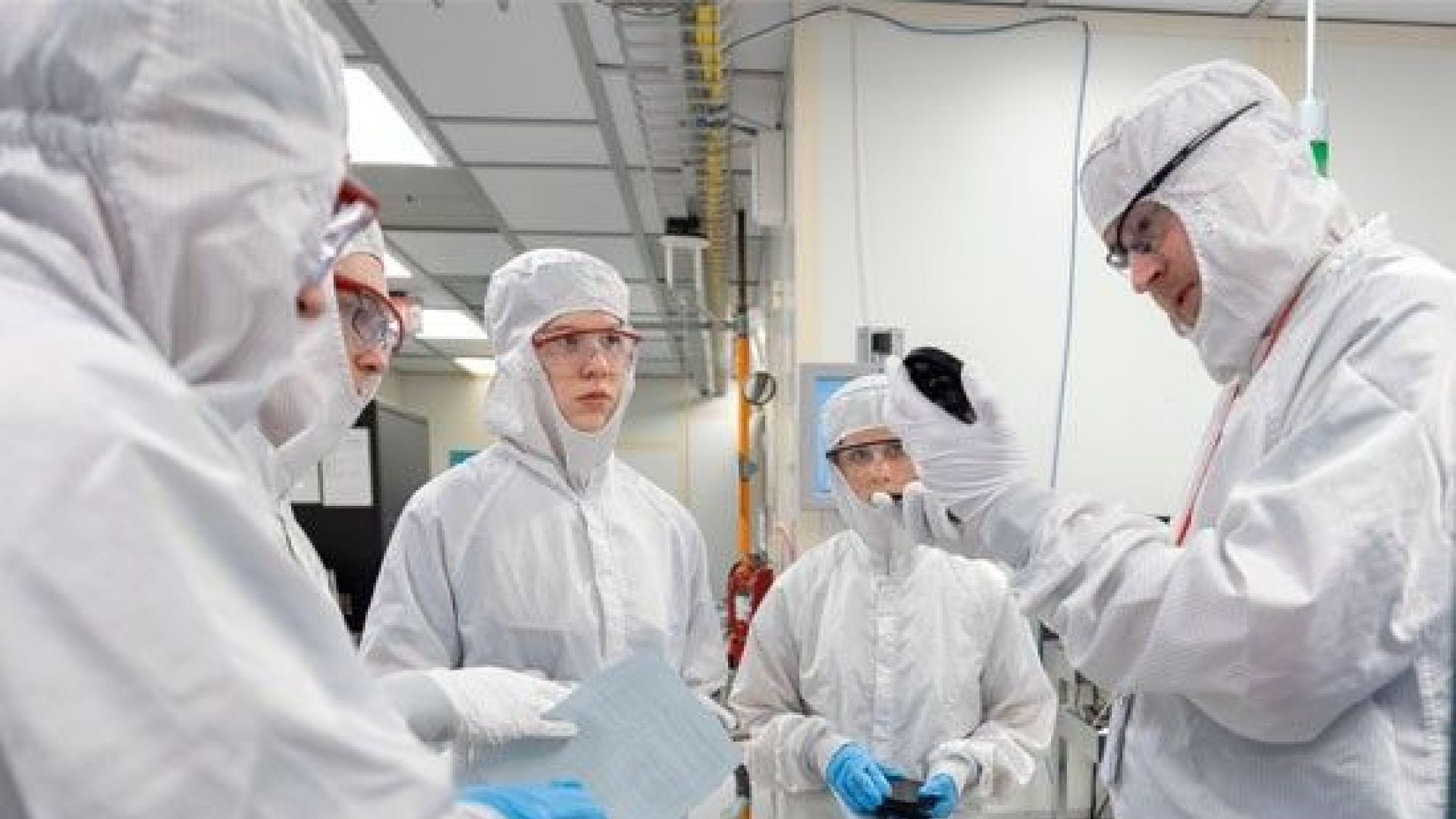 Tom Pennell in clean room Group of people wearing cleanroom suits and safety goggles in a laboratory setting, with one person holding up a device for inspection.